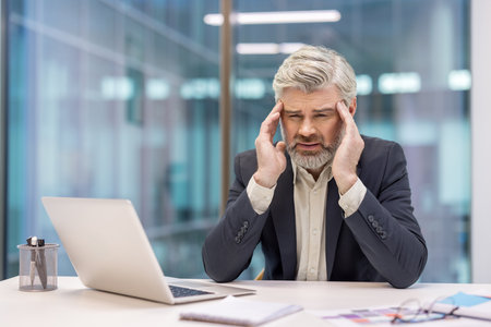 Senior businessman sitting at a desk in a modern office, grappling with a severe headache and expressing physical pain while working on a laptop, indicating burnout and job-related pressureの写真素材