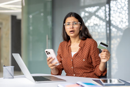 Woman holding a credit card and telephone, looking shocked and experiencing a payment error or fraud issue while trying to make an online purchase using a laptopの写真素材