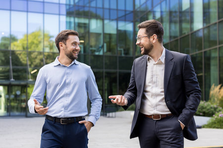 Two smiling adult businessmen walking and having a conversation outside a modern corporate office building, discussing business strategy or partnershipの写真素材