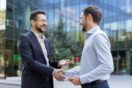 Two smiling professional businessmen are shaking hands, confirming a successful business deal or partnership in an urban setting with a modern office building in the backgroundの写真素材