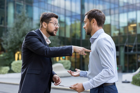 Two angry businessmen are having a tense argument, one pointing a finger while the other gestures in disagreement, depicting workplace stress and conflict outside a modern office buildingの写真素材