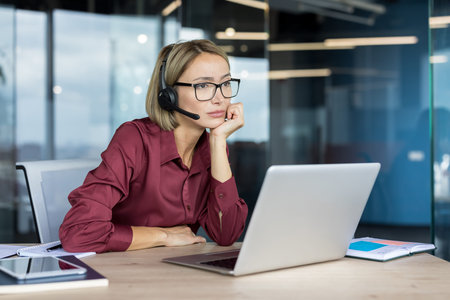 Bored woman with headset working on laptop, providing customer support while looking pensive,...