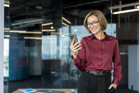 Business professional woman with glasses and short hair standing in a contemporary office, happily engaging with her mobile phone, feeling happy and contentの写真素材