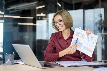 Businesswoman giving a financial presentation during a video call, confidently pointing at a chart while working remotely from a modern office using her laptopの写真素材