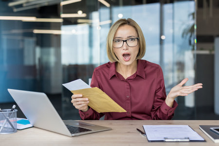 Businesswoman looking shocked and stressed while opening an official envelope with important documents, sitting at a desk in a modern office, reacting to unexpected problems or a big surpriseの写真素材