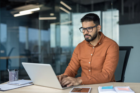Young indian man concentrating on his work, typing on a laptop at a desk in a contemporary office setting, reflecting business productivity and digital technologyの写真素材