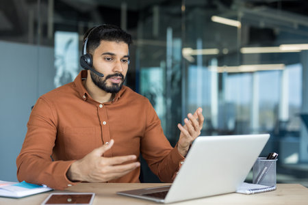 Young bearded man in a brown shirt working in a modern office, having a video conference call on a laptop, actively communicating and gesturing with handsの写真素材