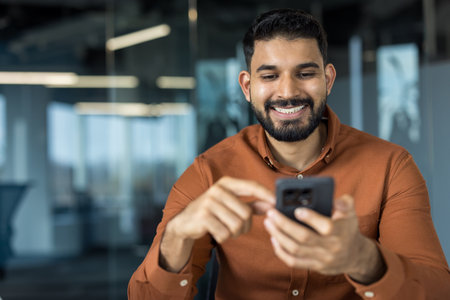 Indian businessman smiling, enjoying connection, holding a mobile phone in a modern office, typing message or checking social media, representing communication and technologyの写真素材