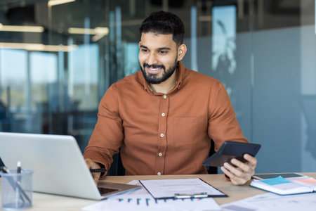 Young indian businessman managing finances and analyzing financial documents while working with a calculator and laptop at a modern office desk, smiling confidentlyの写真素材