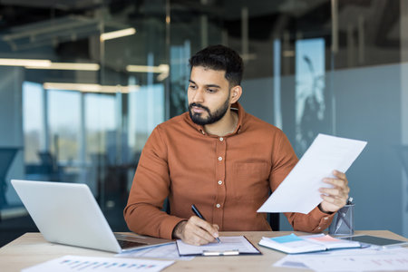 Young man working at a desk, reviewing documents and signing papers while using a laptop in a contemporary office environment, symbolizing business management and professional productivityの写真素材