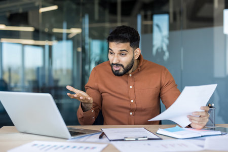Frustrated young businessman confused by difficult work, looking at a laptop and holding paper documents while sitting at an office desk, illustrating concepts of stress and workloadの写真素材
