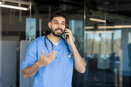 Dedicated male doctor wearing scrubs and a stethoscope engaging in a phone conversation, providing...