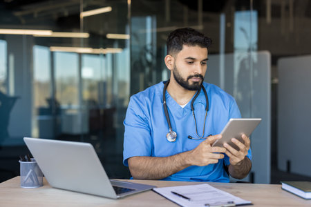 Young male doctor wearing blue scrubs and a stethoscope sitting at a desk, reviewing medical data on...