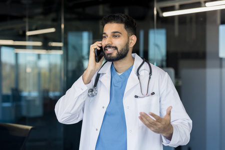 Indian doctor wearing a lab coat and stethoscope is smiling and actively speaking on a mobile phone, representing professional medical communication and telemedicineの写真素材