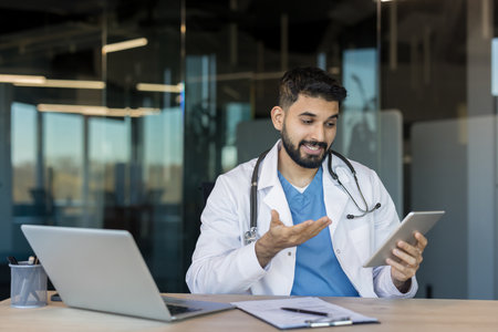 Young male doctor having a telemedicine video call with a patient on a digital tablet, sitting at a desk with a laptop and documents in a modern officeの写真素材