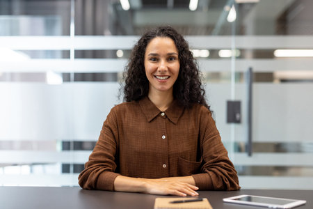 Professional woman with curly hair seated at a dark desk in a modern office, smiling confidently at the camera during a meeting or video call, relaxed and approachable presenceの写真素材