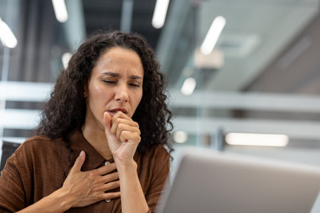 Woman experiencing severe coughing and chest pain in an office environment while working on a laptop, illustrating concepts of illness, sickness, and discomfort during professional obligationsの写真素材