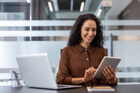 Professional businesswoman with curly hair smiling while using a tablet at a modern desk, multitasking with laptop, showcasing confident remote work, digital connectivity and successの写真素材