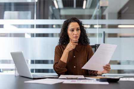 Businesswoman reviewing paperwork and analyzing data, concentrating on important details while working at her desk with a laptop in a professional office settingの写真素材