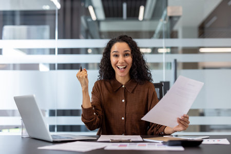 Young mixed race businesswoman celebrating achievement and feeling excited, holding success document and gesturing with a joyful expression while working in a bright office environmentの写真素材