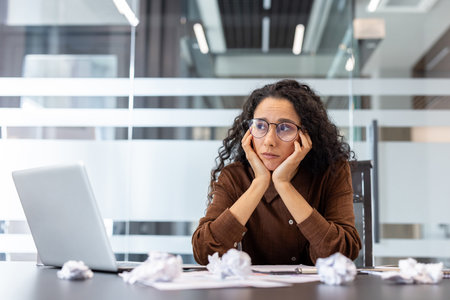 Woman feeling overwhelmed at her office desk, looking frustrated and tired, surrounded by crumpled papers, dealing with work pressure, deadlines, and a lack of inspirationの写真素材