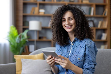 Young woman with curly hair is smiling and looking at camera while holding a digital tablet and...