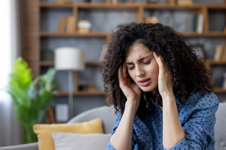 Stressed woman feeling a strong migraine, touching her temples, sitting on the sofa in the living room, suffering from a headache, chronic pain, health problem conceptの写真素材