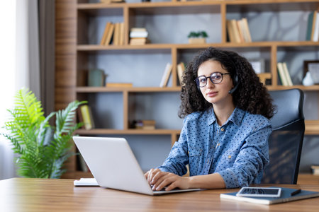 Young woman with curly hair and glasses wearing a headset, focused on laptop at a home office desk providing customer support and virtual assistance during remote workの写真素材