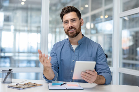 Handsome businessman communicating online, having a virtual meeting, using a digital tablet at a modern office desk, and looking at the camera with a friendly smileの写真素材