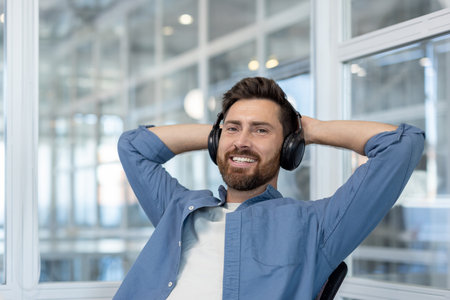 Happy bearded man sitting back in an office chair with hands behind his head, enjoying a break from work while listening to audio to unwind and feel calmの写真素材