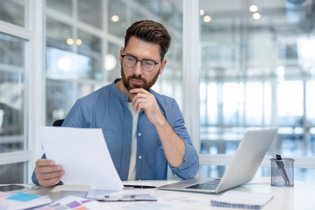 A man in a blue shirt and glasses reviews documents, thoughtfully considering information.の写真素材