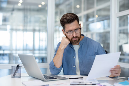 A businessman in glasses reviews paperwork at his desk in a modern office setting, with a laptop and...