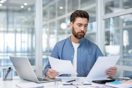 A focused businessman reviews documents at his desk, surrounded by office essentials, captured in a...