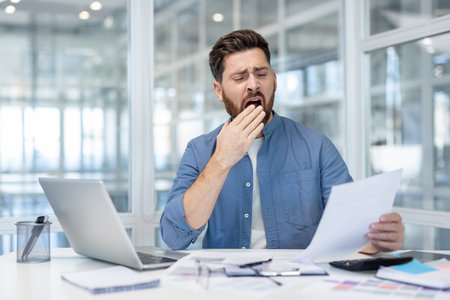 A bored businessman yawns while reviewing documents at his desk, showing signs of fatigue and exhaustion.の写真素材