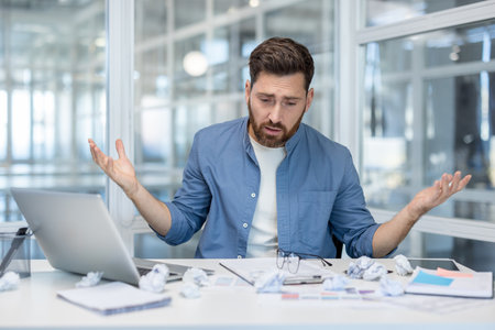 Stressed man sitting at a messy office desk, feeling overwhelmed and confused, raising hands in a gesture of helplessness with crumpled papers around him, experiencing burnout and despairの写真素材