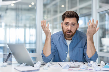 Stressed male employee experiencing burnout at a messy office desk, holding hands up in frustration...