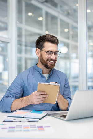 A smiling man with glasses takes notes while looking at his laptop at a modern office.の写真素材