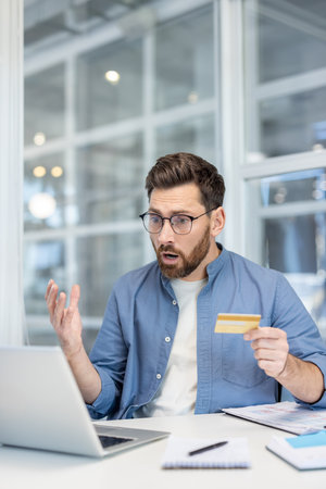 Money transfer error fraud, man got money blocked while working inside office with laptop. Worker upset and worried holding bank credit card in hands.の写真素材