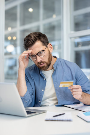 Man finding it difficult to manage debt while holding a credit card and using a laptop for online banking, feeling overwhelmed by financial problems and budget constraintsの写真素材