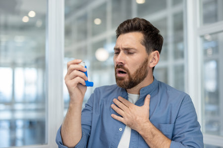 Bearded man having an acute asthma attack, clutching his chest and holding an inhaler, struggling to breathe and appearing anxious and breathless indoors, close-up shotの写真素材