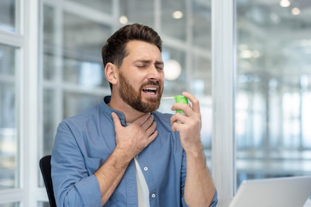 Bearded man at his office desk spraying throat medicine to relieve sore throat and discomfort while...