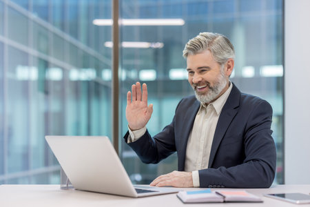 Smiling mature businessman having an online video conference call on a laptop, raising his hand to wave from a bright modern office with large glass windowsの写真素材