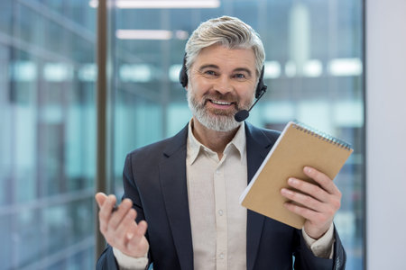 Smiling bearded man wearing a headset with a microphone, holding a notepad and gesturing, providing...