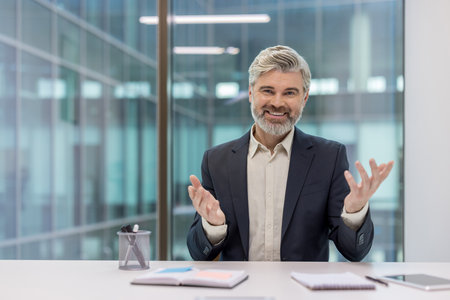 Mature executive, a confident businessman with a grey beard, smiling at the camera and gesturing with his hands during a virtual meeting or online presentation from a modern office deskの写真素材