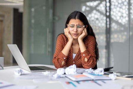 Young woman feeling overwhelmed and frustrated by her work, sitting at a desk with a laptop and...