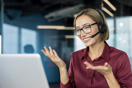 Woman wearing a headset and glasses, smiling and gesturing while working on a laptop, providing professional customer support in a modern call center officeの写真素材