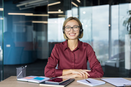 Woman wearing headset and glasses, sitting at an office desk, looking at the camera and smiling, providing online customer service or remote support from a modern workspaceの写真素材