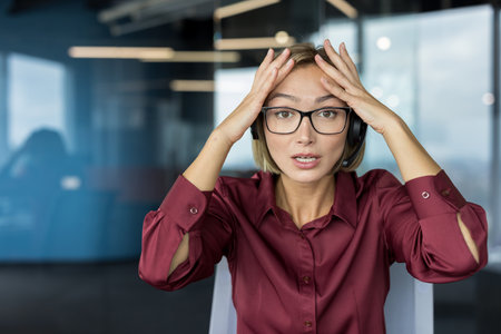 Stressed woman wearing a headset and glasses, holding her head in her hands while looking at the camera, experiencing a problem or mistake at a call center workplaceの写真素材