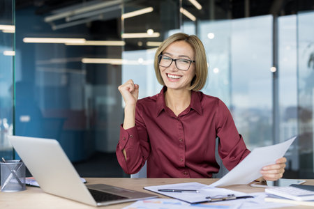 Confident businesswoman celebrating job success, raising fist in victory while holding documents, sitting at a desk with a laptop in a contemporary office environmentの写真素材