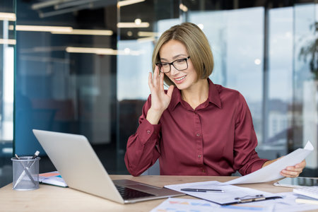 Confident businesswoman smiling and adjusting her glasses while reviewing printed documents at a modern office desk, managing tasks and engaging in productive workの写真素材